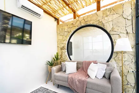 Cozy tropical lounge with gray sofa, pink throw blanket, white pillows, potted plants, stone walls, bamboo ceiling, and large circular window. - Bali Villas